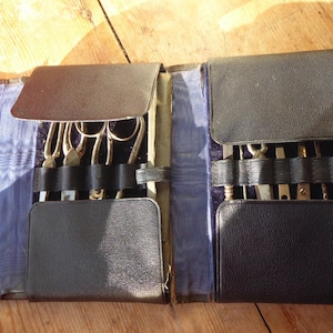 May include: A black leather-effect folding case containing a set of metal tools, including scissors and various implements. The interior features a blue fabric lining with black leather straps to secure the tools in place, and a metal clasp closure.