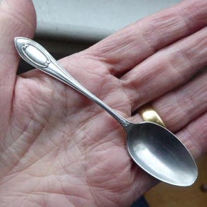 Puede incluir: Una cuchara plateada con un mango ovalado y un cuenco redondeado. La cuchara se sostiene en una mano, mostrando su superficie pulida y su diseño intrincado. La cuchara probablemente se usa para comer o servir.