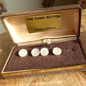 Może przedstawiać: Brązowe pudełko z aksamitu z pozłacanym wykończeniem, zawierające cztery białe perłowe guziki. Pudełko ma złotą etykietę z napisem "The Panic Button" i "Patent Pending."
