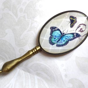 Op de afbeelding: Een antieke handspiegel met een ovaal glas. De spiegel heeft een messing handvat en frame. Het glas toont twee vlinders, een blauwe en een gele, met decoratieve krullen.