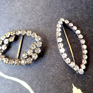 Può includere: Due fibbie vintage con strass. Una è di forma ovale, l'altra a forma di foglia. Entrambe hanno una montatura in metallo argentato e sono incastonate con strass trasparenti. Le fibbie hanno una barra in metallo dorato per l'allacciatura.