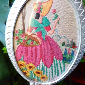 Könnte beinhalten: Ovaler, silberfarbener Handspiegel mit einer gestickten Szene. Die Stickerei zeigt eine Frau in einem rosa Kleid und Strohhut, die eine Blume und einen Korb hält. Gelbe Sonnenblumen und ein grün-weiß gestreifter Hintergrund sind ebenfalls zu sehen.