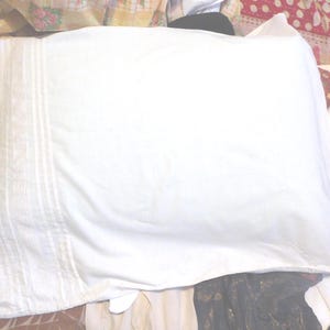 May include: A white pillowcase with vertical stitched detailing on one side. The pillowcase is rectangular and appears to be made of a soft fabric. The background features a mix of patterned fabrics.