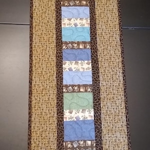 May include: A brown and tan table runner with a brown border and a center strip of blue, green, and white patterned fabric.