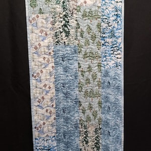 May include: A quilted wall hanging featuring a winter forest scene. The design incorporates various fabrics with snowy trees and blue accents. The rectangular piece is approximately 15 inches wide and 30 inches long.