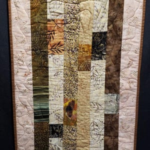 May include: A handcrafted quilt with a rectangular design. The quilt features vertical strips of various earth-toned fabrics, including browns, tans, and greens, with leaf patterns. The border is a light pink with a leaf pattern and a brown edge.