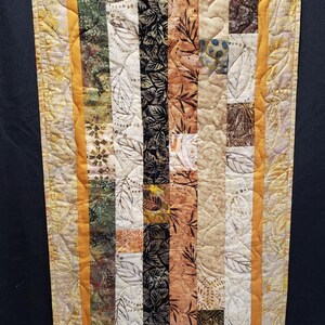 May include: A quilted wall hanging with a rectangular design. The quilt features vertical strips of fabric in various colors and patterns, including leaves and floral prints. The border is a golden yellow with a leaf pattern.