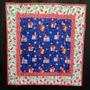 May include: A square, quilted Christmas-themed wall hanging. The center features a blue fabric with repeating images of elves, gifts, and snowflakes. A red dotted border surrounds the center. The outer border is white with red and green Christmas lights.