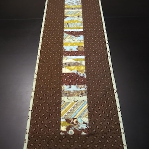 May include: A brown quilted table runner with a central strip of patchwork fabric in shades of brown, blue, and yellow. The patchwork fabric is made up of various patterns and textures.