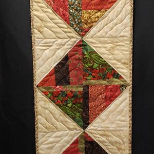 May include: A quilted table runner with a diamond pattern. The diamonds feature various fabrics in red, green, and gold, with floral and abstract designs. The runner is bordered with a cream-colored fabric and a decorative edge.