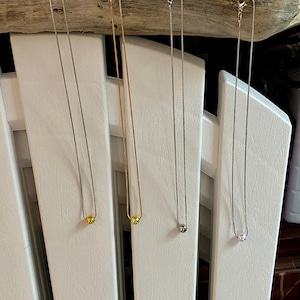 May include: Three necklaces with different colored beads hanging on a white wooden surface. The necklaces are silver, gold, and a darker silver.