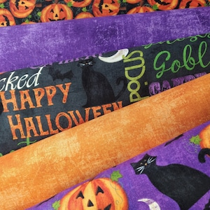 May include: A stack of four different Halloween themed fabric panels. The top panel features a repeating pattern of orange pumpkins with black faces. The second panel is a solid purple color. The third panel is black with white text that reads "Wicked Happy Halloween" and a black cat. The fourth panel is orange with a textured surface. The bottom panel is purple with a repeating pattern of orange pumpkins with black faces and black cats.