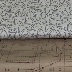May include: A close-up of a beige fabric with a repeating pattern of gray safety pins. The fabric is folded over a brown piece of paper with a sewing pattern printed on it. The pattern includes lines, circles, and the words "Cutting" and "Stitching".