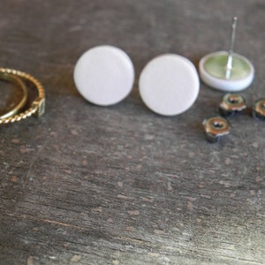 May include: Two white round ceramic earrings with silver posts. The earrings are on a wooden surface with other jewelry pieces.