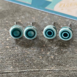 Puede incluir: Cuatro pendientes de cerámica azul turquesa con postes plateados. Cada pendiente tiene un diseño de círculos concéntricos con un centro negro.