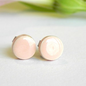 Pode incluir: Um par de pequenos brincos de cerâmica redondos rosa e branco. Os brincos são de cor rosa claro com uma borda branca.
