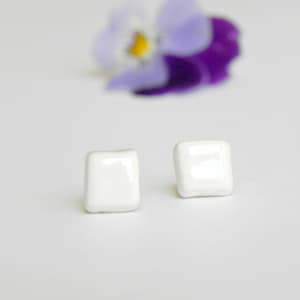 Op de afbeelding: Een paar witte vierkante oorbelletjes van keramiek. De oorbelletjes zijn klein en simpel, met een glanzende afwerking.