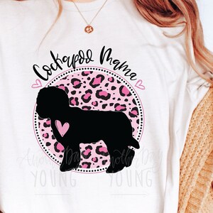 Pode incluir: Uma camiseta branca com um design circular de estampa de leopardo rosa e preto que apresenta uma silhueta preta de um cão Cockapoo. O texto "Cockapoo Mama" está escrito em preto acima do círculo. O texto "Another Day Young" está escrito em preto abaixo do círculo.