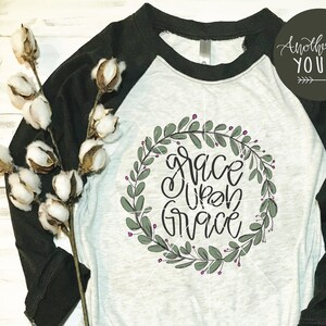 May include: A white and black baseball tee shirt with a green leafy wreath design that says "Grace Upon Grace" in black script. The shirt is lying on a white surface with a cotton boll branch and blue jeans.