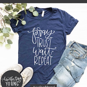 May include: A navy blue t-shirt with white text that reads "pray trust wait repeat". The shirt is laid out on a white surface with a pair of white sneakers and a pair of blue jeans. The text "Another Day Young" is in a circle on a black background.