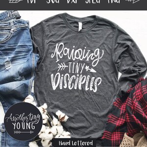 May include: A gray long-sleeved t-shirt with the text "Raising Tiny Disciples" in white. The shirt is laying on a white surface with a pair of blue jeans, cotton bolls, and a plaid blanket.