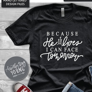 May include: A black t-shirt with white text that reads "Because He Lives I Can Face Tomorrow". The shirt is laid flat on a wooden surface with a pair of red sneakers and blue jeans beside it.