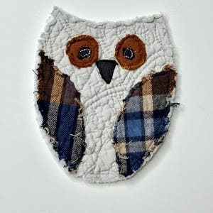 May include: A fabric owl applique featuring a white quilted body with frayed edges. The owl has two brown circular eyes with black centers, a black triangular beak, and two wings made from blue, brown, and tan plaid fabric with raw edges.