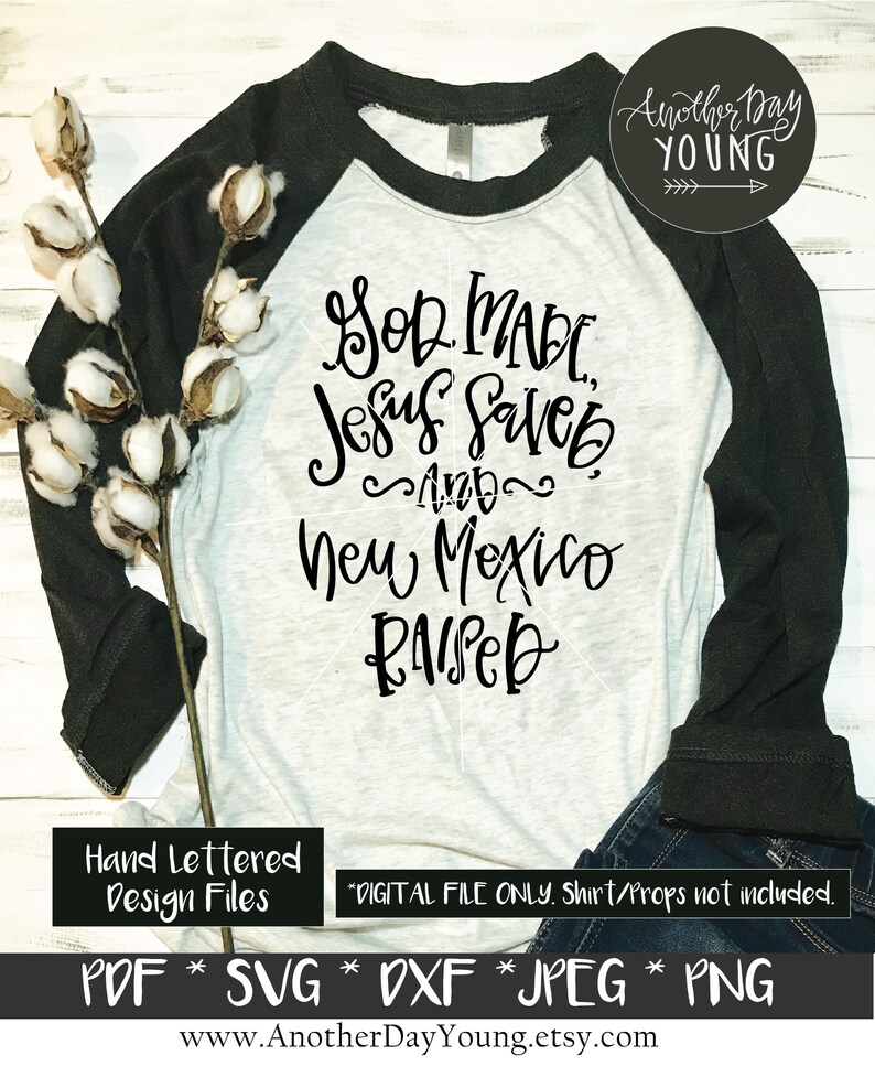 May include: A white t-shirt with black lettering that reads "God Made, Jesus Saved, and New Mexico Raised". The shirt is a baseball style with black sleeves. The image is on a white wood background with a cotton branch.