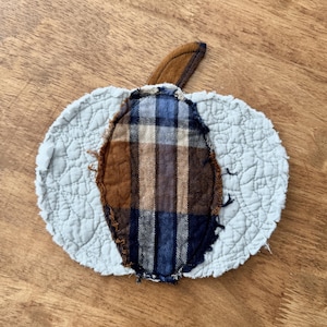 May include: A fabric pumpkin-shaped coaster featuring a light gray quilted background with a central oval patch made of brown, blue, and white plaid flannel fabric. The edges of the plaid patch are frayed, and a small brown fabric stem is attached.