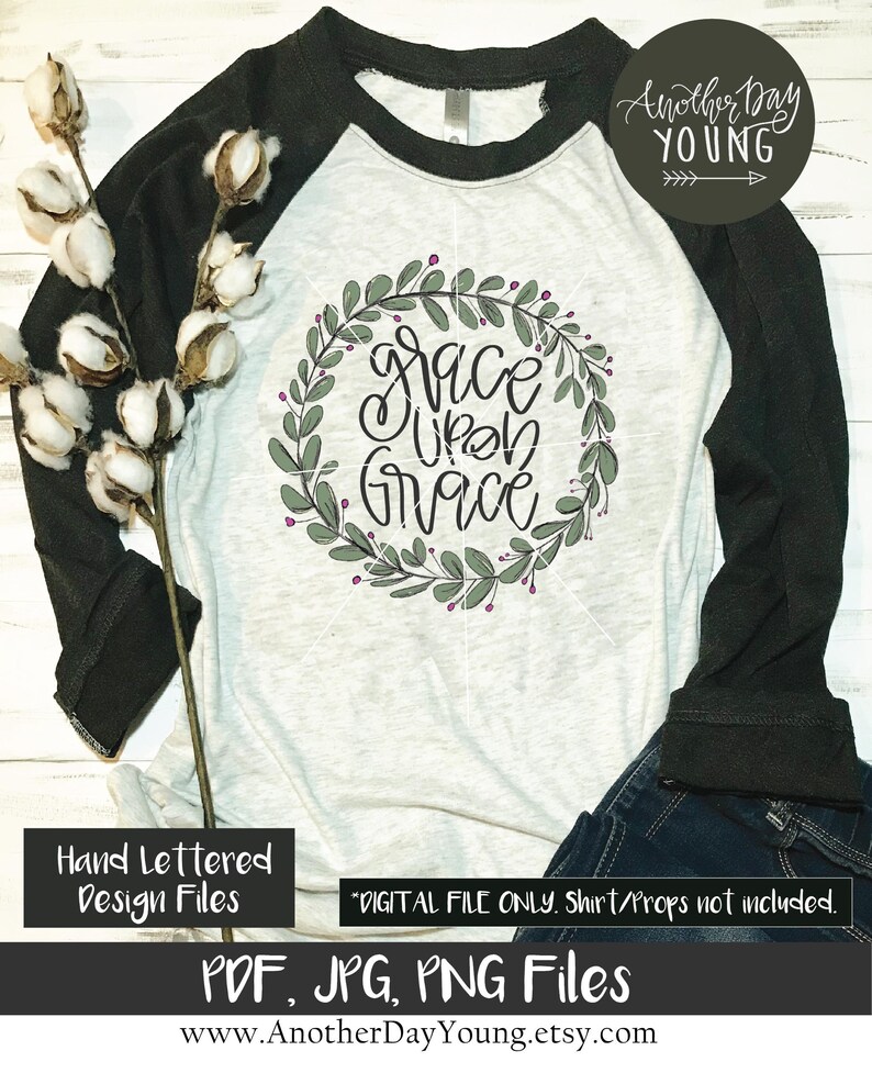 May include: A white and black baseball tee shirt with a green leafy wreath design that says "Grace Upon Grace" in black script. The shirt is lying on a white surface with a cotton boll branch and blue jeans.