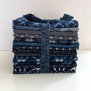 Może przedstawiać: Stos 15 kwadratów tkaniny w kolorze indygo z różnymi wzorami geometrycznymi i kwiatowymi. Tkanina jest związana niebieską wstążką.