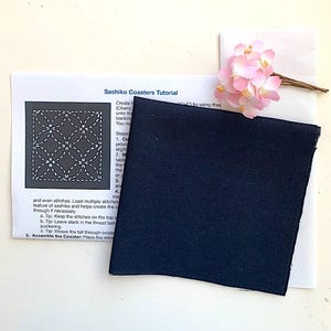 K&ouml;nnte beinhalten: Ein Anleitungsblatt f&uuml;r Sashiko-Untersetzer mit gedrucktem Text und einem Diagramm eines geometrischen Musters, begleitet von einem quadratischen St&uuml;ck dunkelblauem Stoff und einem kleinen dekorativen B&uuml;ndel k&uuml;nstlicher hellrosa Kirschbl&uuml;ten.