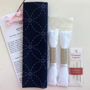 以下が含まれることがあります： 紺色の生地に白い幾何学模様、白い糸2本、針、説明書が入った刺し子刺繍キット。キットにはピンクの花が含まれており、日本の伝統的なステッチング用に設計されています。