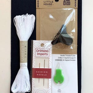 K&ouml;nnte beinhalten: Ein Sashiko-Stickset bestehend aus einem dunkelblauen Stoffquadrat, einem Strang wei&szlig;em Baumwollgarn, einem schwarzen Fingerhut der Marke Little House, zwei Sashiko-Nadeln von Orimono Imports und einem gr&uuml;nen Nadeleinf&auml;dler mit Fadenabschneider.
