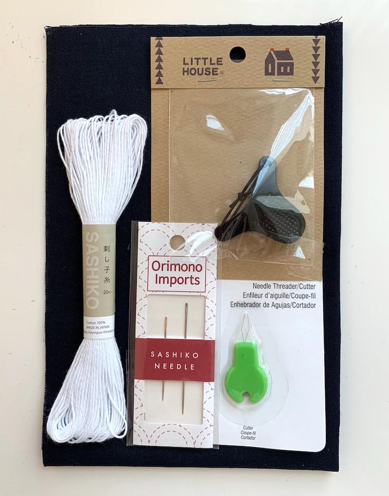 K&ouml;nnte beinhalten: Ein Sashiko-Stickset bestehend aus einem dunkelblauen Stoffquadrat, einem Strang wei&szlig;em Baumwollgarn, einem schwarzen Fingerhut der Marke Little House, zwei Sashiko-Nadeln von Orimono Imports und einem gr&uuml;nen Nadeleinf&auml;dler mit Fadenabschneider.