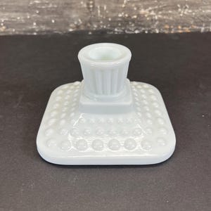 May include: A white, square candle holder with a textured, dotted base and a cylindrical top. The candle holder is made of opaque glass and has a classic, vintage design. The top has vertical ridges.