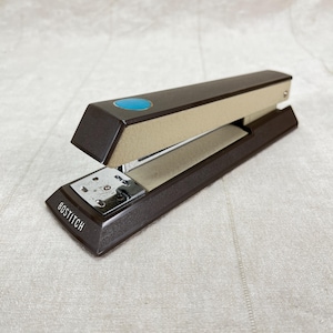 May include: A brown and beige Bostitch stapler with a blue accent button. The stapler has a metal base and the brand name is printed on the front. The stapler is open, ready for use.