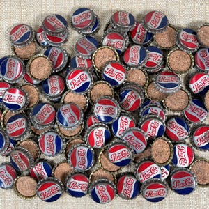 May include: A collection of vintage Pepsi Cola bottle caps. The caps feature a red, white, and blue design with the Pepsi Cola logo. The caps are made of metal and have a cork backing. The caps are scattered on a beige surface.