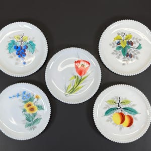 May include: Five white ceramic plates with beaded edges, each featuring a different hand-painted fruit or floral design. The designs include grapes, a tulip, blackberries, sunflowers, and peaches. The plates are arranged on a dark surface.