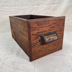 May include: A rustic wooden drawer with a dark brown finish. The drawer features a decorative metal handle with a label holder. The wood grain is visible, adding to the vintage aesthetic. Ideal for storage or display.