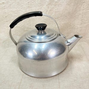 May include: A silver-colored kettle with a black handle and lid knob. The kettle has a rounded body, a spout, and a metal handle that arches over the top. The handle has a black plastic grip. The lid knob is also black.