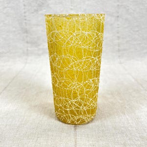 May include: A tall, yellow glass with a white, scribbled pattern. The glass has a cylindrical shape and is made of a translucent material. The white pattern covers the entire surface of the glass, creating a textured effect. The glass is likely used for drinking.