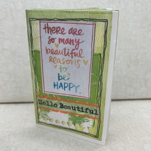 Può includere: Carta verde con una citazione colorata che recita "Ci sono così tante belle ragioni per essere felici." La carta ha anche il testo "Hello Beautiful" in nero.