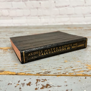 May include: A vintage book titled "Audels Mathematics and Calculations for Mechanics." The book has a black cover with gold lettering and a reddish-brown edge. The book is lying on a weathered, light-colored wooden surface.