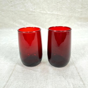 May include: Two red-tinted, stemless drinking glasses. The glasses have a rounded, cylindrical shape with a dark red base that fades to a brighter red at the rim. The glasses are set against a neutral background.