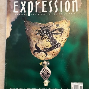 May include: A magazine cover for Expression, a magazine about creative arts. The cover features a mermaid illustration on a piece of parchment with a silver charm hanging from it. The magazine title is in large white letters and the subtitle is "Sharing the Spirit of Creative Arts". The cover also includes the text "Cords of Clay, Renaissance Angel, Piano Hinge Journal, Menorah Men, Colored Pencils 101, Spinning in Circles".
