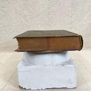 May include: An antique book with a dark cover and aged, yellowed pages. The book's spine is visible, showing a gold-colored edge. It rests on a white, weathered pedestal, suggesting a vintage or historical theme.