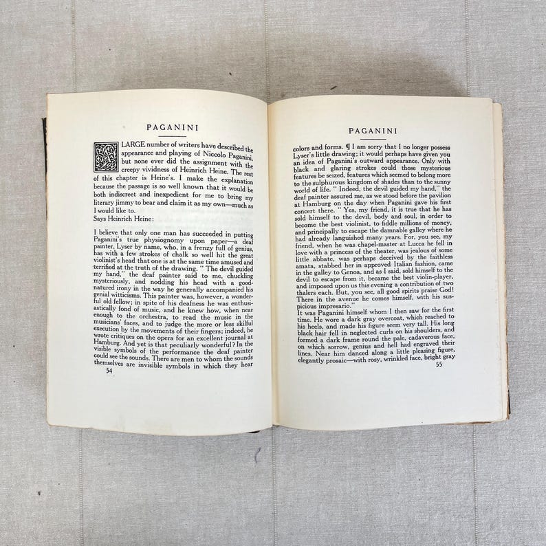 May include: Open book pages with text about Paganini, featuring a decorative initial. The text is in English, with a focus on the appearance and playing of Niccolo Paganini. The pages are aged, with a light beige tone.