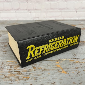 May include: A black book titled "AUDELS REFRIGERATION and AIR CONDITIONING GUIDE" with yellow lettering. The book has a textured cover and light-colored pages. The book is on a weathered wooden surface.