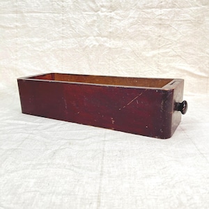 May include: A rectangular wooden drawer with a dark reddish-brown finish. The drawer has a small, round, black knob on one end. The interior of the drawer is a lighter brown. The drawer sits on a white, textured surface.